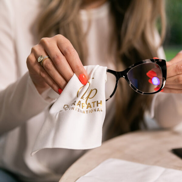 Czyścik Sunbeam Optic Wipe VIP — użycie, producent Sunbeam Optic Wipe VIP: biały czyścik ze złotym nadrukiem w dłoni przy okularach — oryginalne zdjęcie producenta.